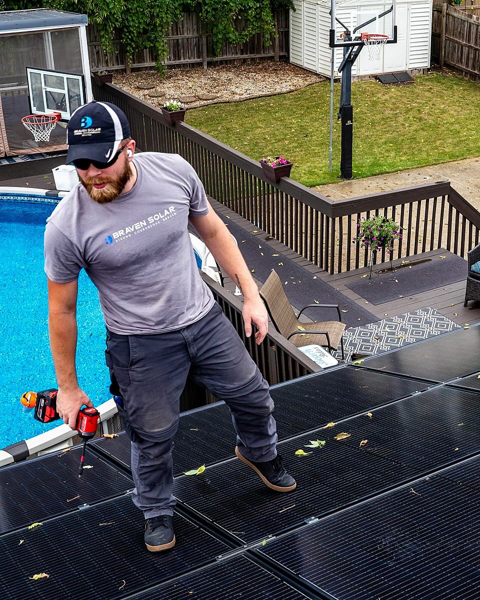 Aerial view of a Braven installer walking a rooftop solar array with an impact driver, a backyard pool below