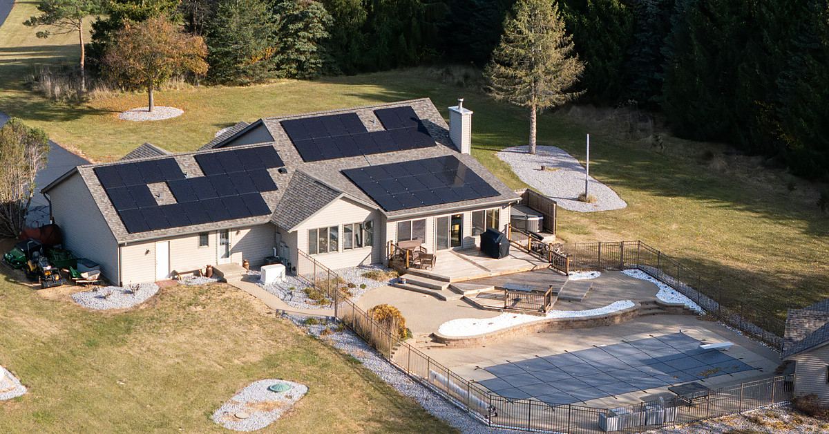 Aerial view of a rural ranch home with solar arrays across three roof faces and mature trees behind