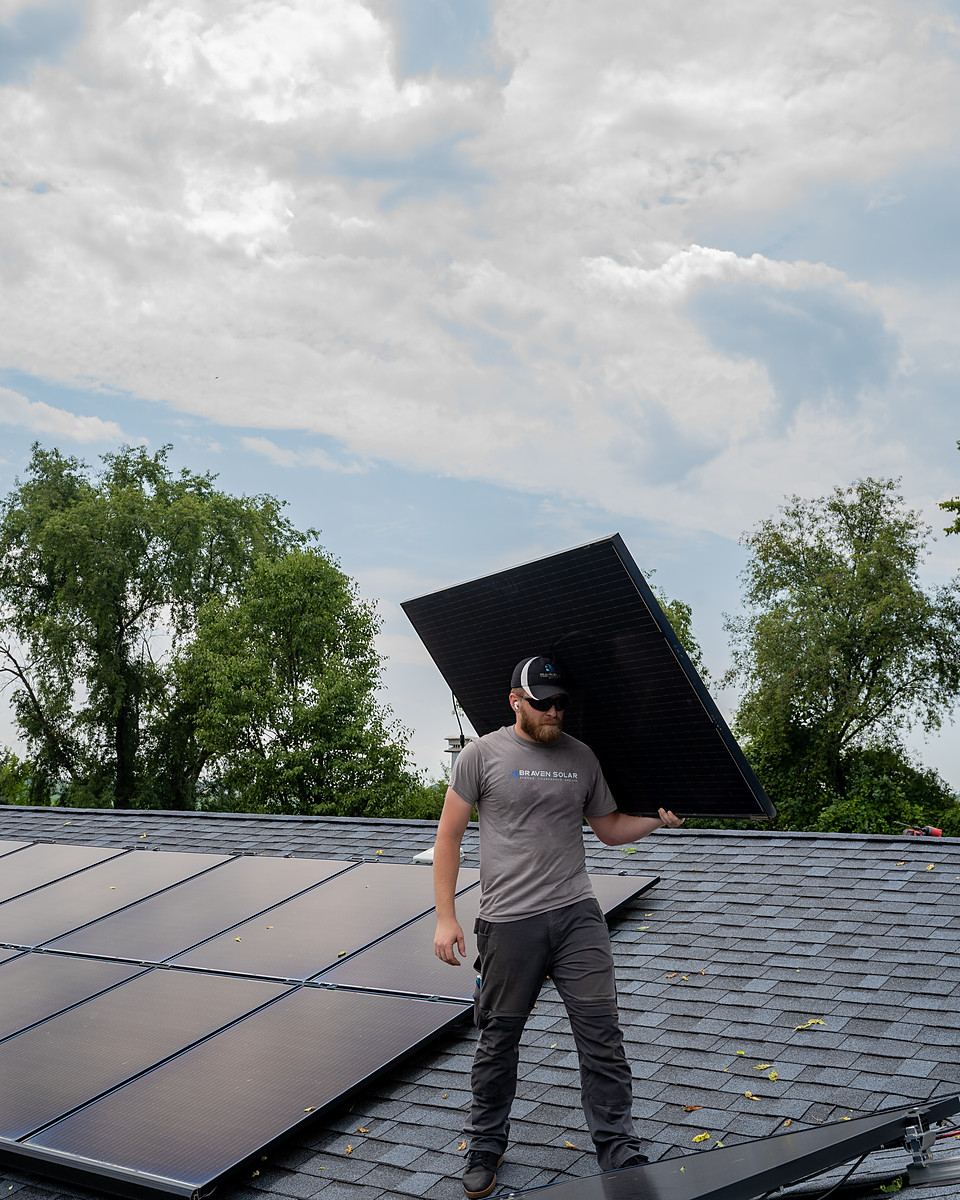 A Braven installer carrying a solar module across a shingle roof against the sky