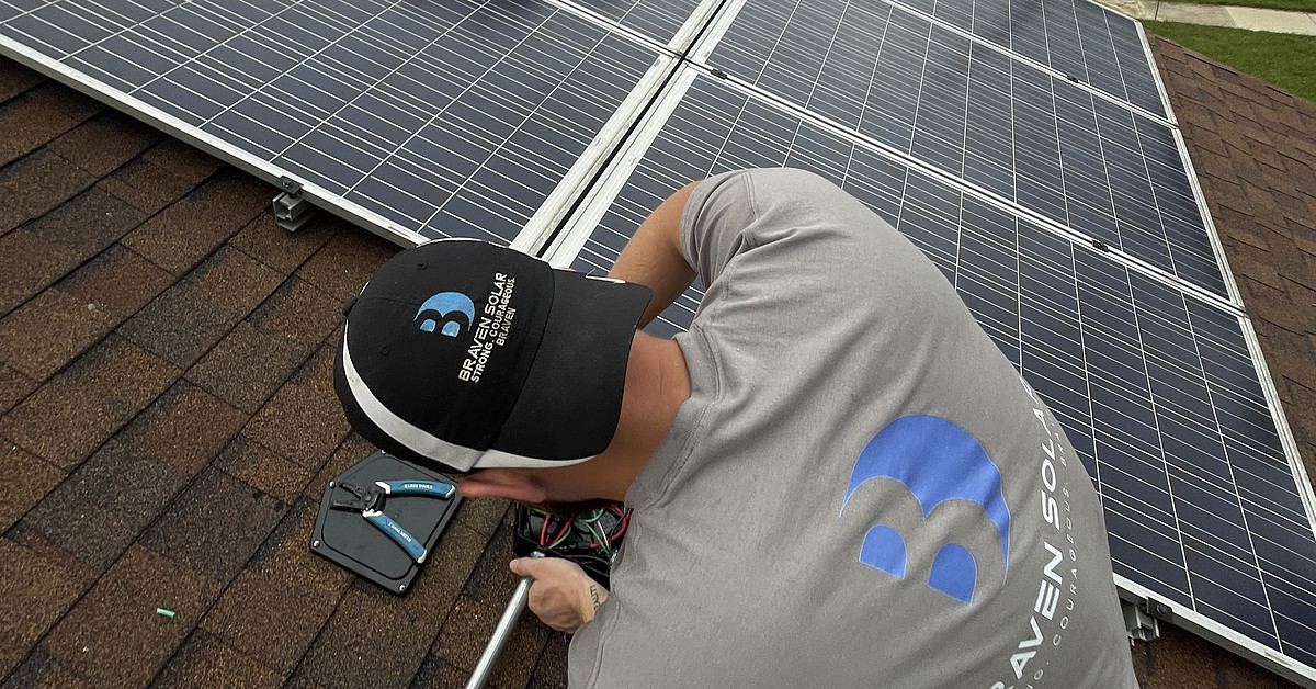 Installer kneeling on a roof beside a solar array with the street behind