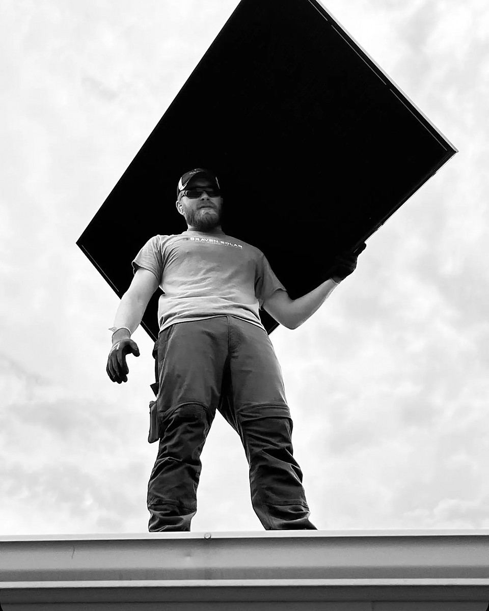 Black-and-white photograph of a Braven installer on a roofline lifting a solar module overhead